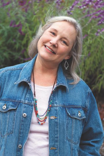 Katherine Applegate author photo. A white woman with shoulder length white-blonde hair wearing a jean jacket, a white t-shirt, and a beaded necklace stands and softly smiles with her head slightly tilted to the right.