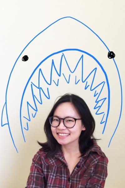 Remy Lai author photo. An Asian woman wearing glasses and a red plaid shirt in front of a line drawing of a blue shark with its mouth open around her head.