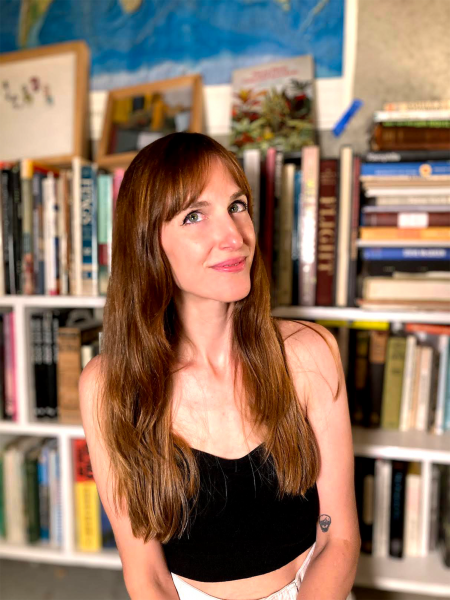 Leah Hayes photo. A white woman with long straight brown hair stands in front of a bookshelf in a black tank top and white pants.